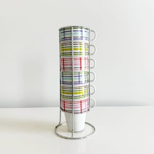May include: A set of six stacked coffee cups with a colorful plaid pattern in a chrome wire rack. The cups have a white interior and a handle. The rack is circular at the base and top, with vertical supports.