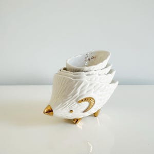 May include: A set of four white ceramic measuring cups stacked to resemble a bird. The bird has gold accents on the beak, feet, and a curved design. The smallest cup reads "1/4 Cup".
