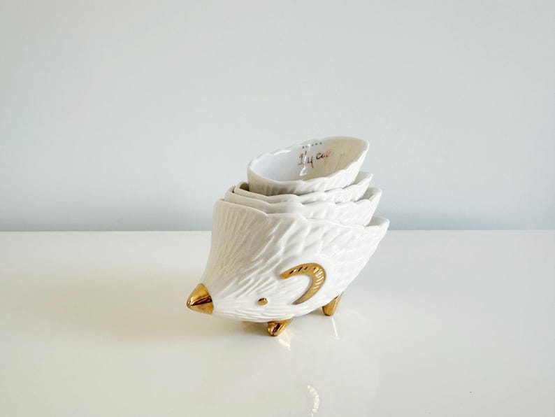 May include: A set of four white ceramic measuring cups stacked to resemble a bird. The bird has gold accents on the beak, feet, and a curved design. The smallest cup reads "1/4 Cup".