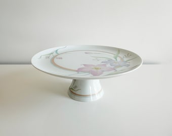 May include: White cake stand with a floral design featuring pink and purple flowers. The stand has a round base and a raised platform.