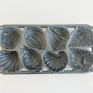 May include: A gray metal baking mold with eight seashell-shaped cavities. The mold features detailed designs of various shells, including scallops and conch shells. The rectangular mold is made of a heavy metal.