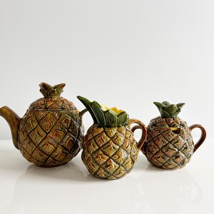 May include: A set of three ceramic pineapple-shaped kitchenware items. The set includes a teapot, creamer, and sugar bowl. The items are decorated with a brown and green pineapple design.