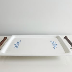 May include: A rectangular white serving tray with blue floral designs and dark brown wooden handles. The tray is made of ceramic and has a glossy finish. The handles are attached with silver metal hardware. A decorative piece for serving food.