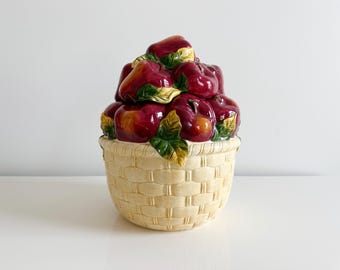 Op de afbeelding: Een keramische koekjespot in de vorm van een mand vol met rode appels. De mand is geweven en heeft een geelbruine kleur. De appels hebben groene bladeren en zijn realistisch geschilderd.