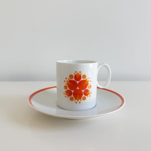 May include: White ceramic mug and saucer set. The mug features a vibrant orange floral design. The saucer has a white surface with an orange rim. The set is designed for serving hot beverages.