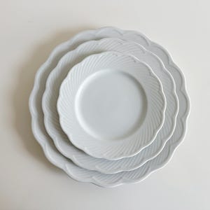 May include: Three white ceramic plates with a scalloped edge and a raised, swirling design. The plates are stacked on top of each other.