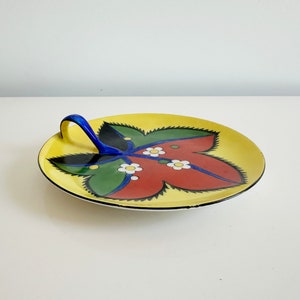 May include: A yellow ceramic serving dish with a blue handle. The dish is decorated with a stylized red, green, and black leaf design with white flowers.