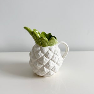 May include: White ceramic pitcher shaped like a pineapple with green leaves on top.