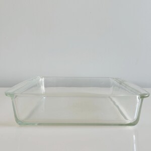 May include: A clear, rectangular glass baking dish with slightly curved edges. The dish is empty and sits on a white surface, reflecting the light. The overall design is simple and functional, suitable for baking and serving.