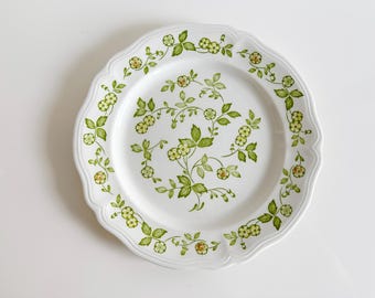 May include: A white ceramic dinner plate with a green and yellow floral pattern. The plate has a scalloped edge.