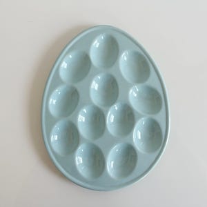 May include: A light blue ceramic egg tray shaped like a large egg, featuring twelve individual oval indentations arranged in a symmetrical pattern for holding eggs. The surface has a smooth, glossy finish with a raised outer rim border.