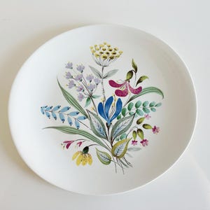May include: A round white ceramic plate featuring a central botanical illustration of a wildflower bouquet. The design includes yellow, blue, purple, and pink flowers with green leaves and stems painted in a detailed, artistic style.