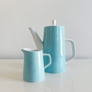 May include: A light blue ceramic coffee pot with a white spout and handle, and a matching creamer. The coffee pot has a white lid. The set is displayed on a white surface.