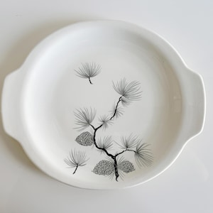 May include: A white ceramic serving dish with two handles. The dish features a black and gray illustration of a pine branch with pine cones. The design is centered on the dish.