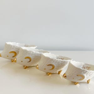 May include: Four white ceramic measuring cups with gold accents, shaped like hedgehogs. Each cup has a gold crescent moon design and gold feet. The text "1/4 cup" is visible on the rim of each cup.