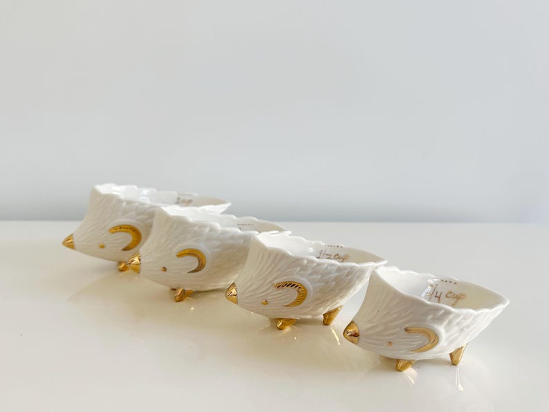 May include: Four white ceramic measuring cups with gold accents, shaped like hedgehogs. Each cup has a gold crescent moon design and gold feet. The text "1/4 cup" is visible on the rim of each cup.