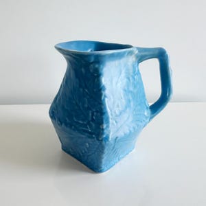 May include: A blue ceramic pitcher with a square base and a handle. The pitcher has a textured leaf pattern and a wide opening. The color is a vibrant, solid blue. The pitcher is likely used for serving beverages.