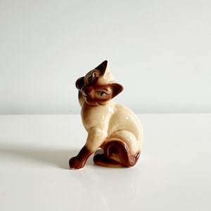 May include: A ceramic Siamese cat figurine in a seated pose. The cat has a cream-colored body with dark brown markings on its face, ears, paws, and tail. The figurine is detailed with a glossy finish.