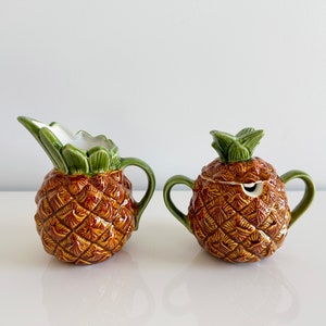 May include: A pineapple-shaped ceramic creamer and sugar bowl set. The creamer has a handle and a spout. The sugar bowl has a lid and two handles. Both pieces are decorated with green leaves and brown pineapple skin.