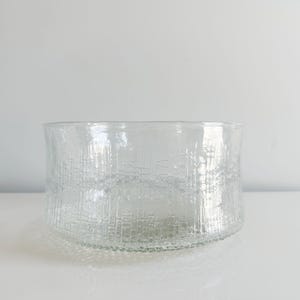 May include: A clear glass bowl with a textured, bark-like pattern. The bowl has a wide, rounded shape and sits on a white surface. The glass appears thick and sturdy, with a slightly irregular rim.