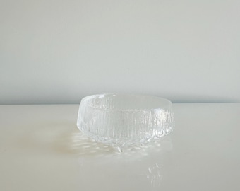 May include: A clear glass bowl featuring a textured exterior with vertical ridges and a smooth interior. The bowl has a wide, circular opening and is supported by three small, integrated glass feet at the base, creating a raised profile.