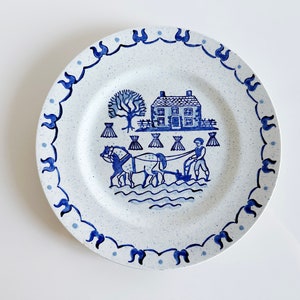 May include: Blue and white ceramic dinner plate with a farmhouse scene featuring a house, a tree, a farmer, a horse, and a plow. The plate has a blue and white scalloped edge.