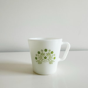 May include: White ceramic mug with a green floral design. The design features stylized leaves and flowers.