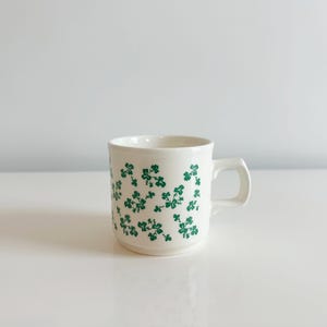 May include: A white ceramic mug featuring a repeating pattern of small green shamrocks across the exterior surface. The mug has a cylindrical body, a flat base, and a sturdy side handle, providing a simple and functional design for daily use.