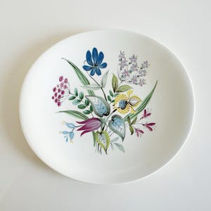 May include: A white ceramic plate with a floral design. The design features a bouquet of flowers in shades of blue, purple, yellow, and green. The plate has a round shape and a smooth surface.