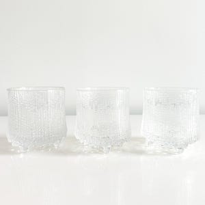 May include: Three clear, textured glass tumblers with a unique bark-like design. The glasses are cylindrical with a slightly flared base and are displayed on a white surface. The glasses are ideal for serving beverages.