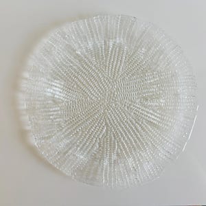 May include: A clear glass charger plate with a textured, radial pattern. The plate is round and has a slightly raised edge.