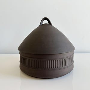May include: A dark brown ceramic lidded container. The lid is conical with a small handle. The base has a band of vertical lines. The container sits on a white surface.
