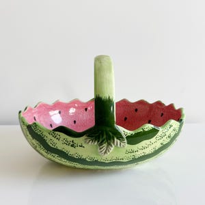 May include: A ceramic watermelon-shaped basket with a handle. The interior is pink with black seeds, and the exterior is green with dark green stripes. The handle is light green with a dark green base and a leaf accent.