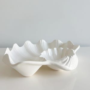 May include: A white ceramic serving dish in the shape of a clam shell. The dish has a wavy, textured design and is divided into two sections. The dish is on a white surface.