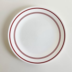 May include: A white ceramic dinner plate with a red rim and two red lines around the center.