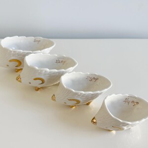 May include: Set of four white ceramic measuring cups with gold accents. Each cup is shaped like a hedgehog and labeled with measurements: 1 cup, 1/2 cup, 1/3 cup, and 1/4 cup. The cups have a textured exterior and gold feet.