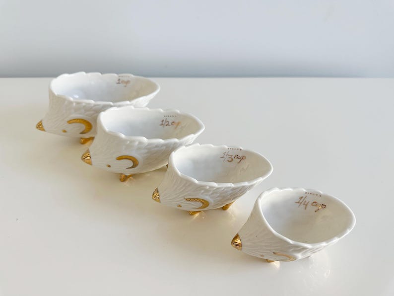 May include: Set of four white ceramic measuring cups with gold accents. Each cup is shaped like a hedgehog and labeled with measurements: 1 cup, 1/2 cup, 1/3 cup, and 1/4 cup. The cups have a textured exterior and gold feet.