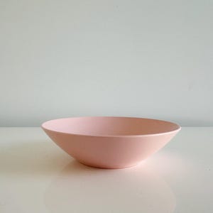 May include: A pale pink bowl with a wide, shallow design. The bowl has a smooth, matte finish and sits on a white surface. The bowl's shape is a simple, elegant cone.