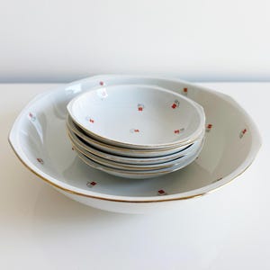 May include: A set of white ceramic serving dishes. The set includes a large, shallow bowl and a stack of smaller bowls. Each piece is decorated with a gold rim and a repeating pattern of red and gray squares.