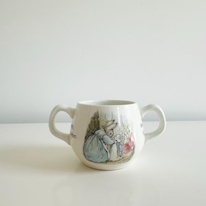 May include: A white ceramic two-handled mug featuring a colorful illustration of a mother rabbit in a blue dress tending to a smaller rabbit. The text "Peter" is printed on the side of the mug. The mug has a rounded body and two side handles.