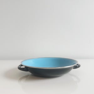 May include: A black enamel pan with a light blue interior and two small handles. The rim is silver. The pan is sitting on a white surface.