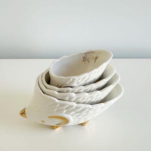 May include: A set of four white ceramic measuring cups stacked together, resembling a hedgehog. The cups have a textured surface and gold accents on the feet and snout. The top cup has gold text that reads "1/4 cup".
