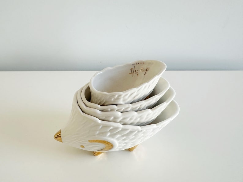 May include: A set of four white ceramic measuring cups stacked together, resembling a hedgehog. The cups have a textured surface and gold accents on the feet and snout. The top cup has gold text that reads "1/4 cup".