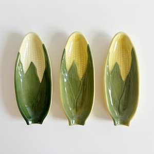 May include: Three ceramic corn-shaped serving dishes. Each dish features a yellow corn cob design with green husks. The dishes are oval-shaped and rest on a white surface. The design is a charming addition to any kitchen.