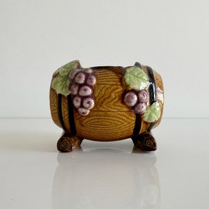 May include: A ceramic planter shaped like a brown wooden barrel with black metal bands. The barrel is decorated with green leaves and purple grapes.
