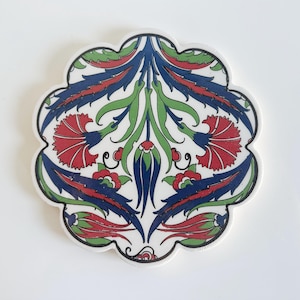 May include: A white ceramic coaster with a scalloped edge, featuring a symmetrical floral design. The design includes red, blue, and green stylized flowers and leaves. The coaster is likely used for placing drinks on a table.