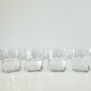 May include: Four clear, textured drinking glasses with a unique crumpled design. The glasses are arranged in a row on a white surface, reflecting the light. The glasses are made of clear glass.