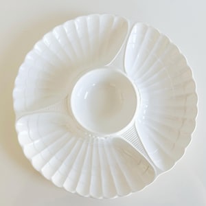 May include: A white ceramic serving platter shaped like a large scallop shell, featuring three distinct outer compartments and a central circular bowl. The surface has a ribbed, fan-like texture radiating from the center to the scalloped edges.