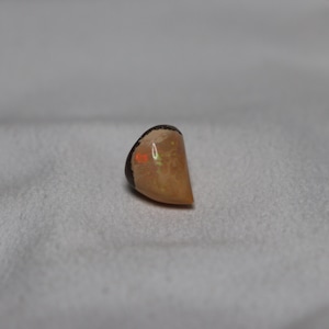 May include: A light brown, teardrop-shaped opal gemstone with a dark brown backing. The opal has a slight iridescent sheen.
