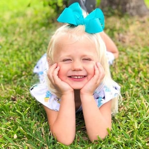 May include: A young child with blonde hair smiles while lying in the grass. The child wears a white dress with a blue floral pattern and a large turquoise bow in their hair. The child rests their chin on their hands.